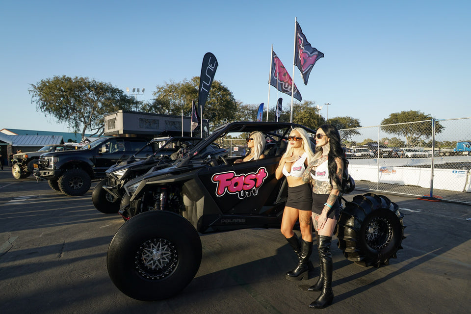 Vista lateral de un UTV negro de FastUTV con modelos junto a la puerta y banderas sobre el techo